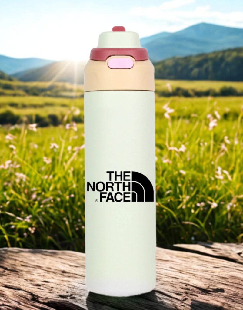 A light-colored North Face water bottle standing upright on a wooden surface outdoors, with a sunny green field, wildflowers, and distant mountains in the background.