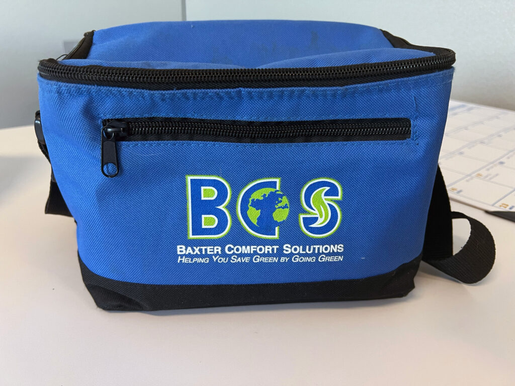 A photo of a blue lunch cooler with a zipper closure with a green and white logo on the front from Baxter Comfort Solutions.