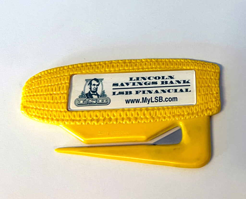 Photo of a letter opener in the shape of a yellow ear of corn with a white and green logo from Lincoln Savings Bank.