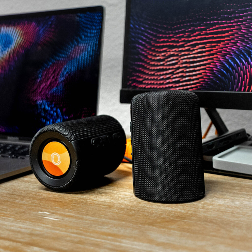 A pair of black breakaway wireless speakers sits on a wooden desk between a laptop and monitor displaying vibrant digital patterns. One speaker stands upright while the other lies on its side, showing a circular orange logo on the end.