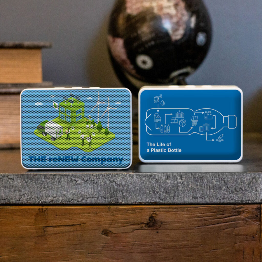 Two rectangular wireless speakers with custom eco-themed designs sit on a wooden surface in front of stacked books and a globe. One speaker shows an illustrated green energy scene with people, wind turbines, and a recycling truck, labeled “THE r-NEW Company.” The other displays a blue infographic titled “The Life of a Plastic Bottle,” showing the recycling process from bottle to new product.