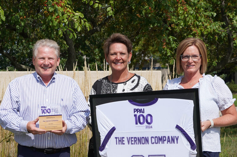 Three people are standing outdoors in front of a green leafy background. The person on the left is holding a wooden plaque that reads 'PPAI 100 The Vernon Company.' The person in the middle is wearing a black and white patterned top. The person on the right is holding a framed white and purple sports jersey with the text 'PPAI 100 THE VERNON COMPANY' printed on it.