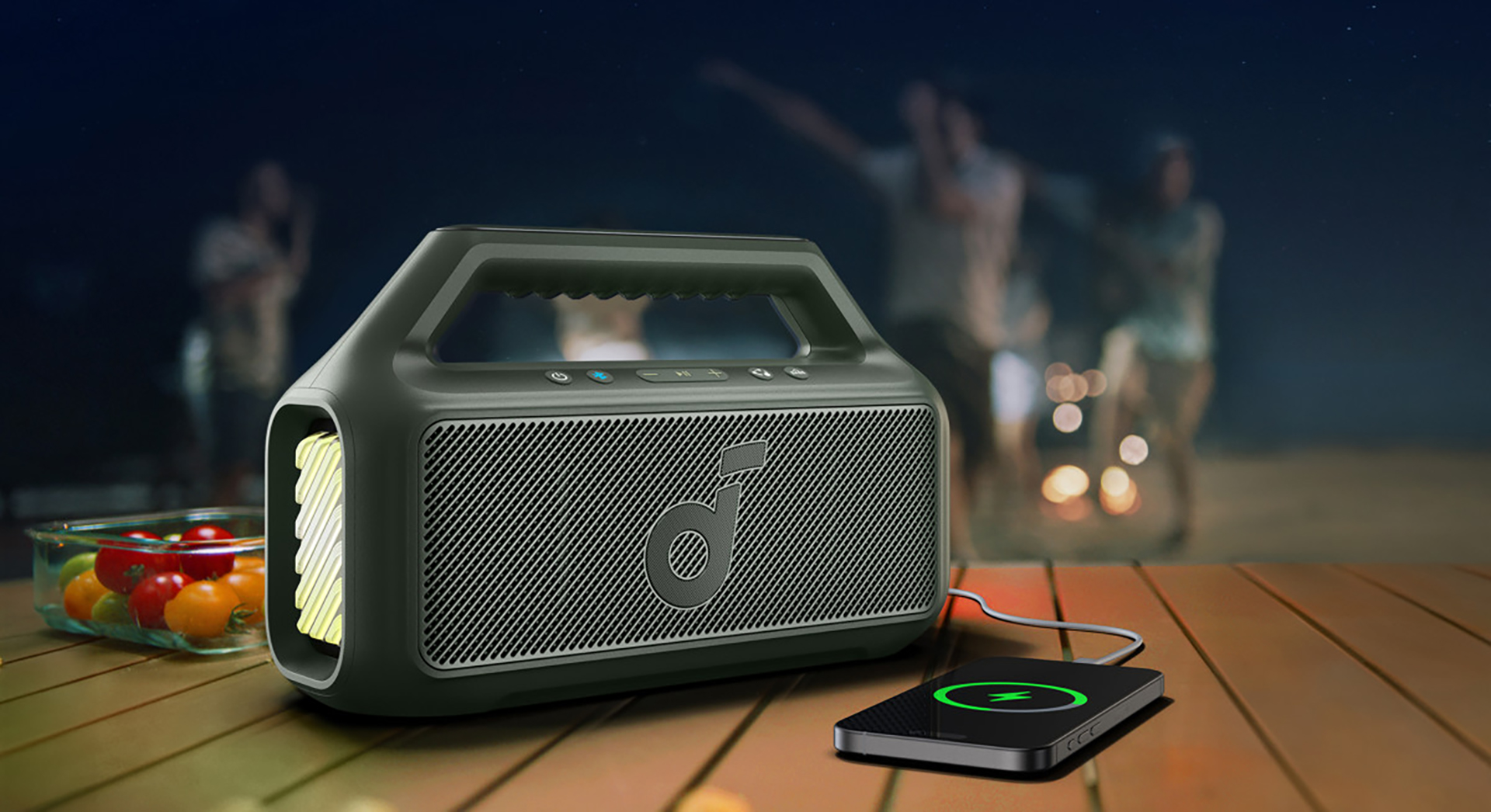 A Black Anker Soundcore Boom speaker and power bank with a yellow light, charging a smartphone on a picnic table, as people in the background enjoy an outdoor event at night.