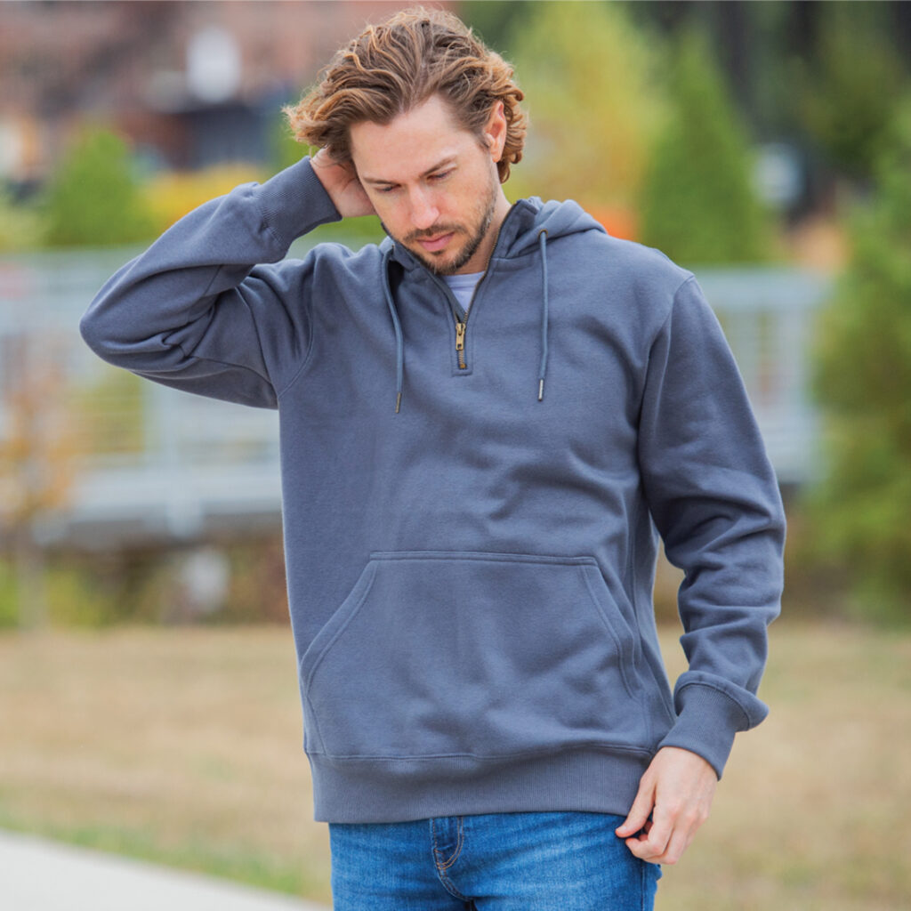 Person in gray hoodie and jeans standing outdoors with greenery in the background.