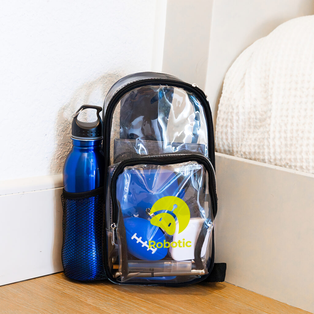 A clear backpack with black trim and a front pocket displaying the logo 'Robotic' in yellow and green. The backpack contains various items, including a blue water bottle in the side mesh pocket, a blue stress ball shaped like an American football and other small objects. The backpack is placed on a wooden floor next to a white bed with a textured blanket.