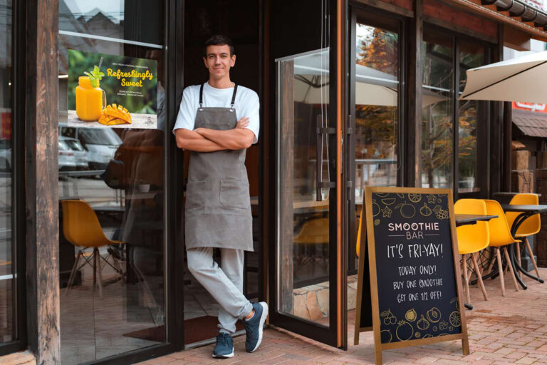 Person in apron stands outside a smoothie bar with a chalkboard sign offering a smoothie deal and a poster showing orange smoothies.