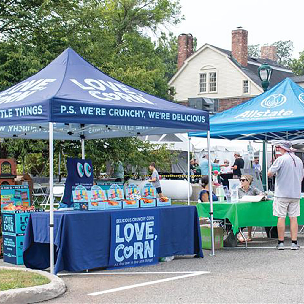 Outdoor market scene with two blue tents—one for 'LOVE, CORN' displaying snack packages, and one for Allstate with people gathered around. A large house and trees are in the background.