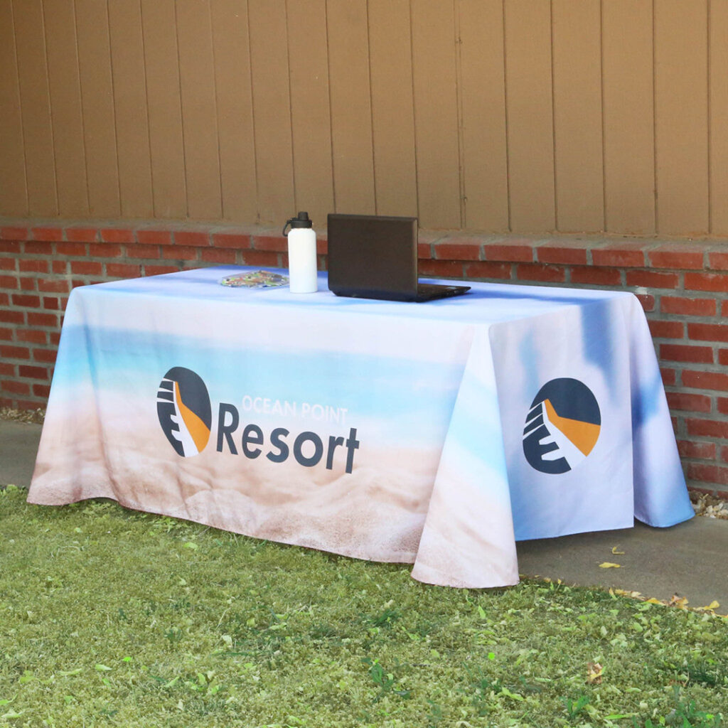 Outdoor table with 'Ocean Point Resort' branding, displaying a laptop, white water bottle, and brochures, set against a brick and wood-paneled wall.