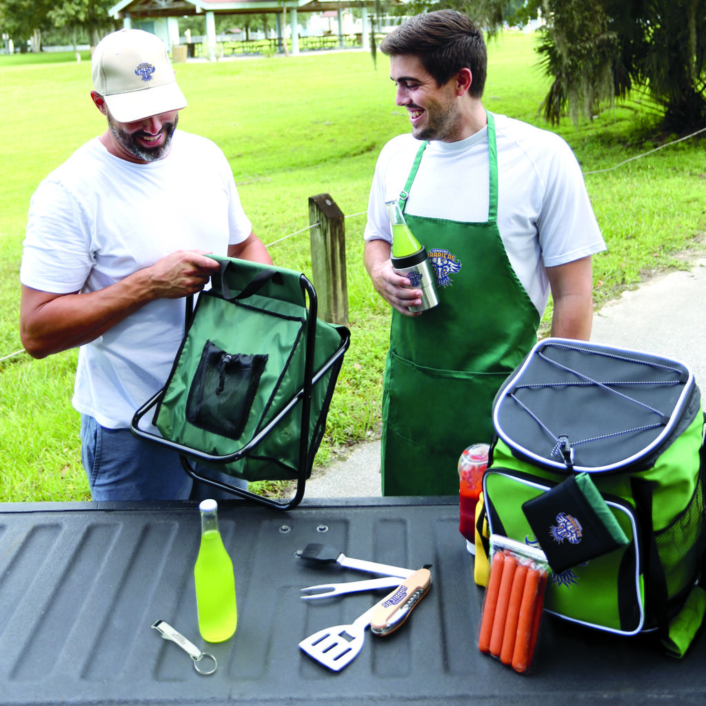 Two men are standing outdoors near the tailgate of a pickup truck. One man is wearing a white t-shirt and blue jeans, holding a green cooler bag seat. The other man is wearing a white t-shirt with a branded green apron over it, holding a bottle of yellow drink that is inside an insulated can cooler. On the tailgate in front of them are various items including branded green cooler bag, hot dogs, grilling utensils (spatula and tongs) and bottles of drinks. The background shows an open grassy area with trees and a pavilion structure.