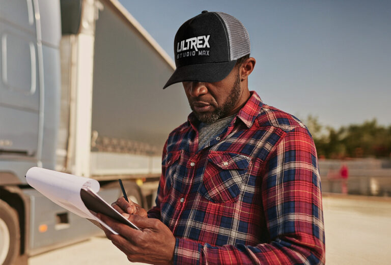 Person in plaid shirt and 'ULTREX' cap holding a clipboard in front of a truck.