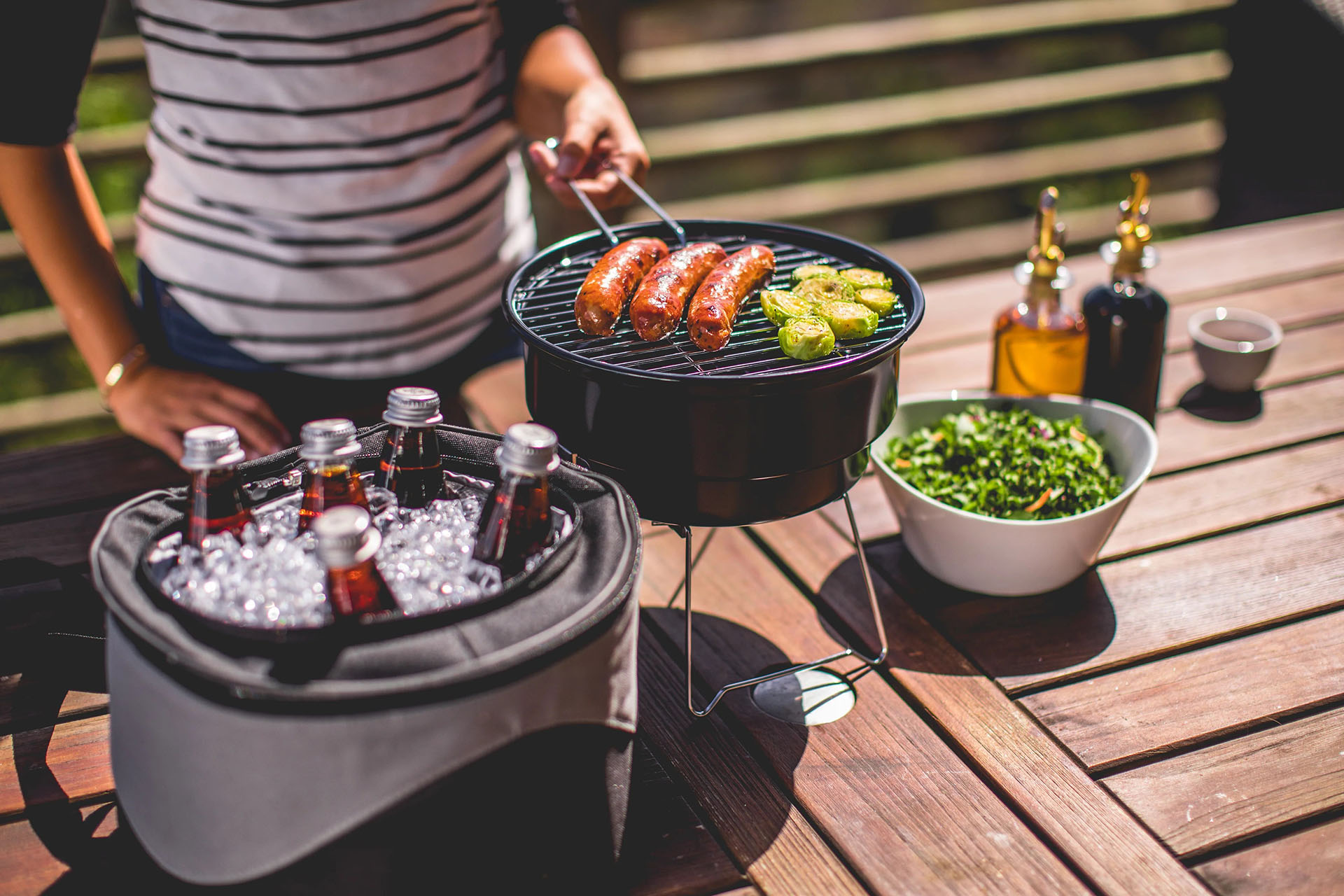 A person is grilling sausages and vegetables on a small round charcoal grill placed on a wooden table. The person is holding tongs and wearing a striped shirt. On the table, there is also a cooler filled with ice and bottled drinks, a bowl of salad, two bottles of condiments or oils, and a small cup.
