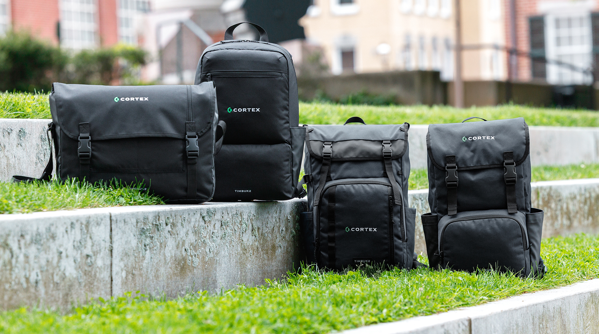 Four black Cortex bags, including backpacks and messenger bags, displayed on concrete steps with grass.