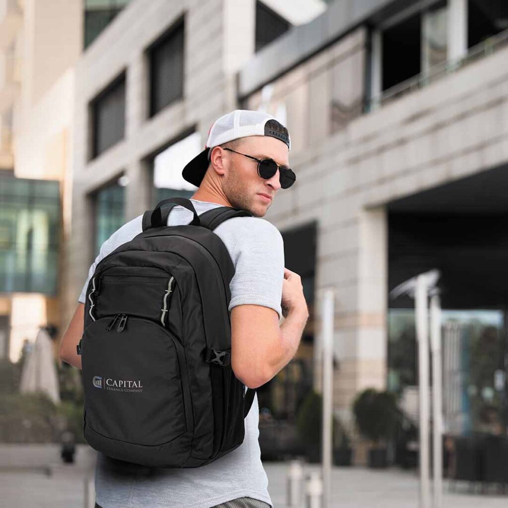 Person in grey t-shirt and white cap carrying a black "CAPITAL" backpack in front of a modern building with windows and greenery.