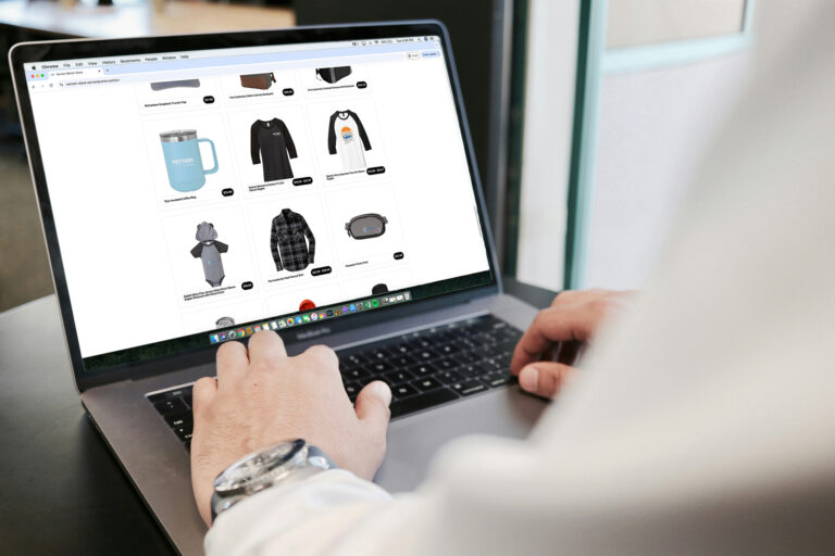 A person is browsing an online store on a laptop. The screen shows items like a blue mug, black hoodie, long-sleeve shirt with a colorful design, gray onesie with ears, plaid shirt, and an oval-shaped item. The person wears a watch and has hands on the keyboard.