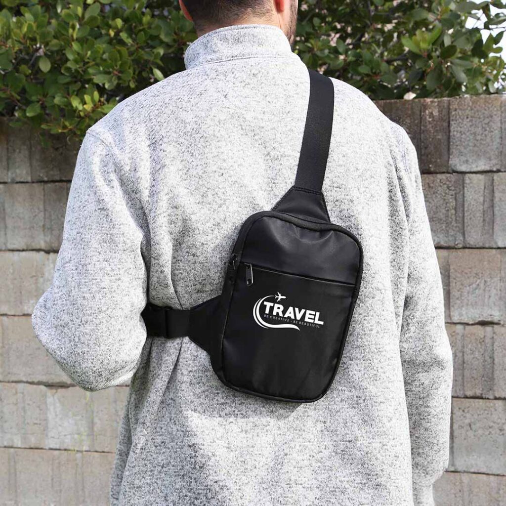 Person in gray fleece jacket with a black "TRAVEL" crossbody bag standing in front of a concrete wall with greenery.
