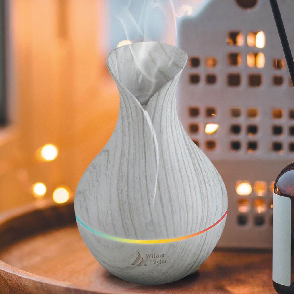 Wood grain essential oil diffuser with colorful LED light ring emitting mist, branded “Wilson Taylor,” set against warm lights and a decorative lantern.