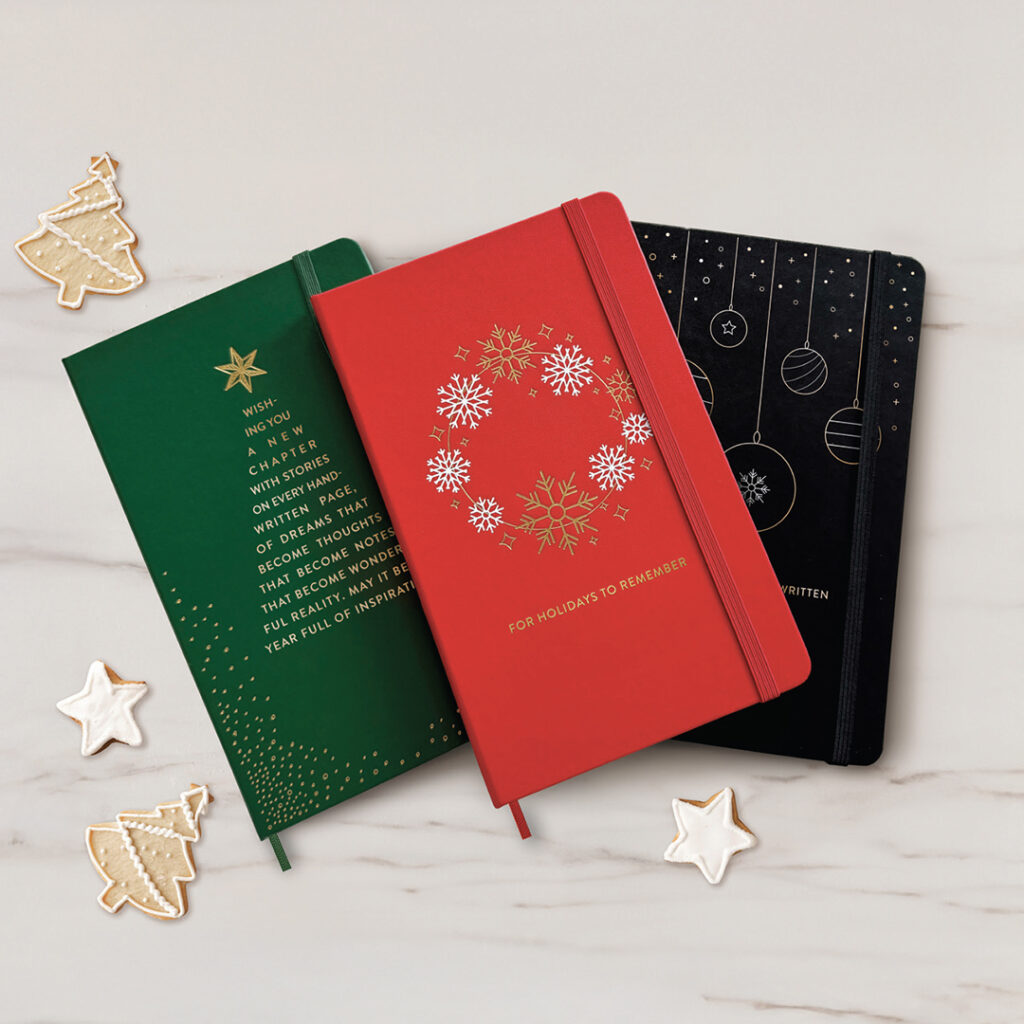 Three festive notebooks with holiday designs and cookies on a light surface.