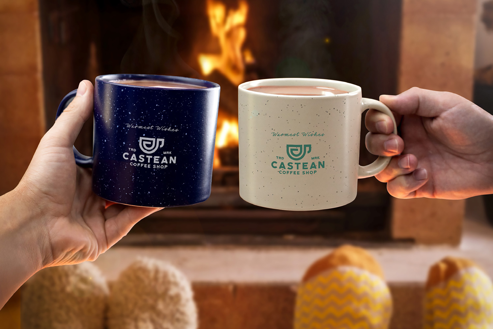 Hands holding speckled coffee mugs with “Warmest Wishes” in front of a cozy fireplace and pillows.