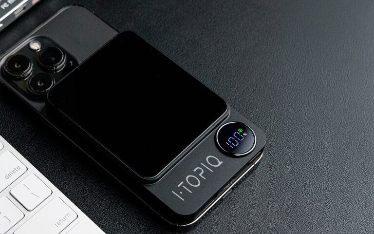 Black iPhone with triple camera and iTOPIQ wireless charger displaying 100% battery, placed on a black surface beside a white keyboard.