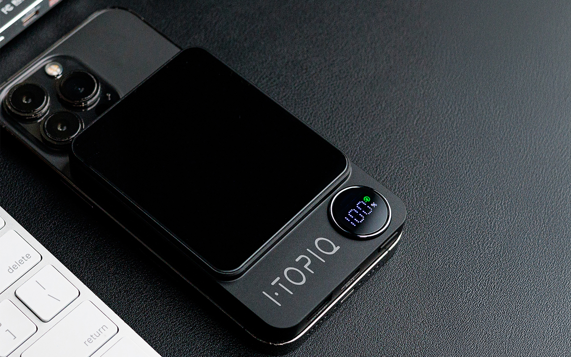 Black iPhone with triple camera and iTOPIQ wireless charger displaying 100% battery, placed on a black surface beside a white keyboard.