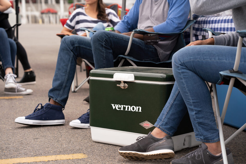 Outdoor gathering with casually dressed people seated on folding chairs around a green and white "Vernon" cooler.