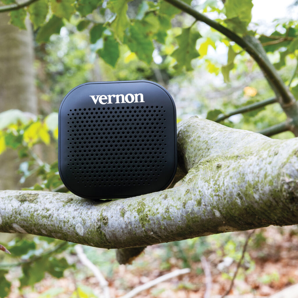 Compact black Vernon-branded speaker with rounded edges placed on a tree branch, surrounded by green foliage in an outdoor setting.