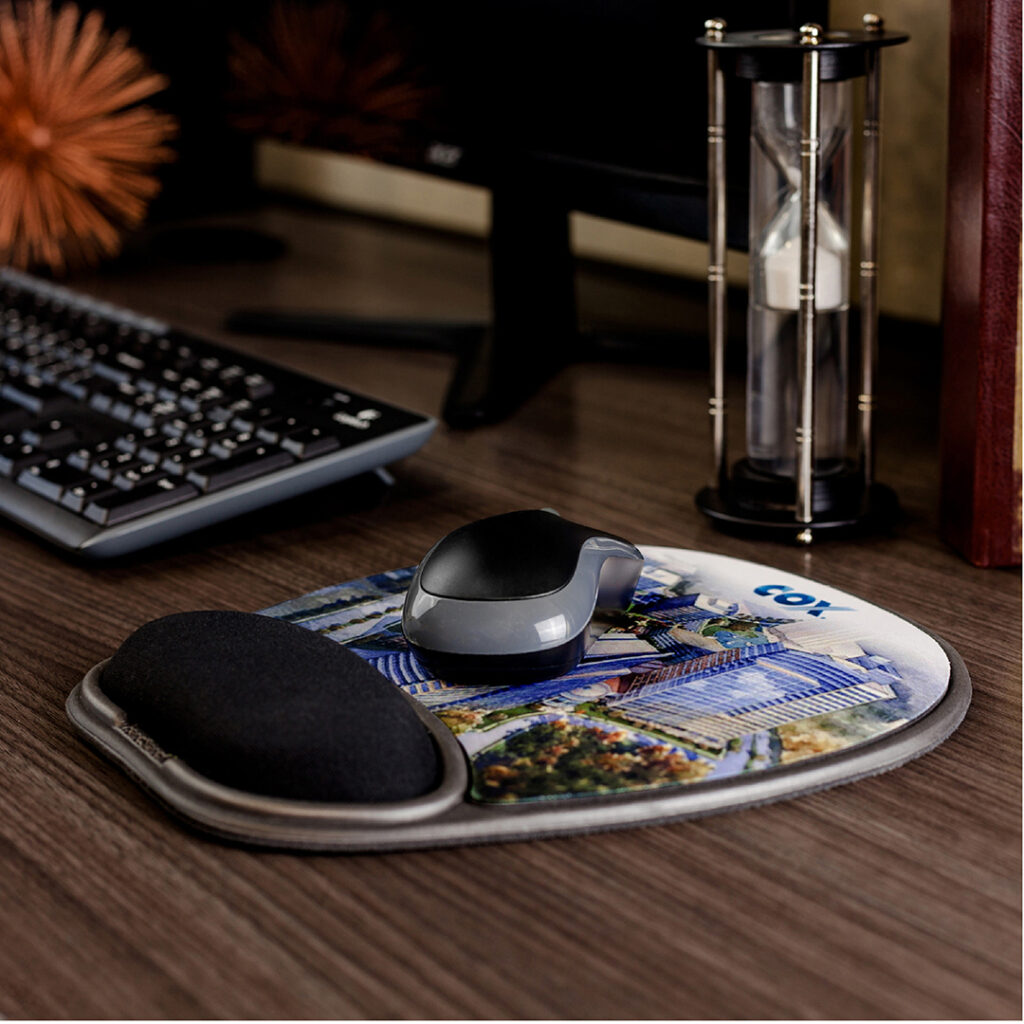 Computer mouse on a custom printed mouse pad with wrist support, next to a keyboard and an hourglass on a wooden desk.