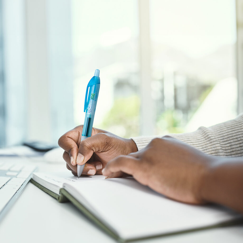 Close-up of hands writing in a notebook with a blue pen at a desk near a computer keyboard.