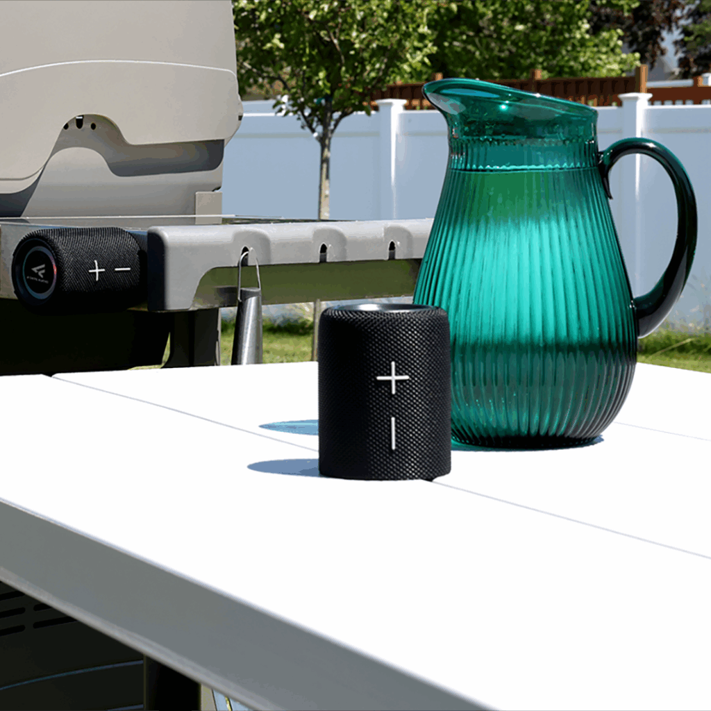 Black portable Bluetooth speaker and green glass pitcher on a white outdoor table near a grill in a backyard setting.
