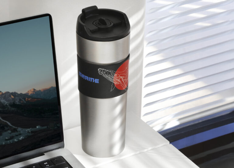Stainless steel travel tumbler with a black flip-top lid sits on a white desk beside an open laptop, featuring a black wrap with a blue “MARINE” imprint and a red whale graphic, lit by sunlight through window blinds.