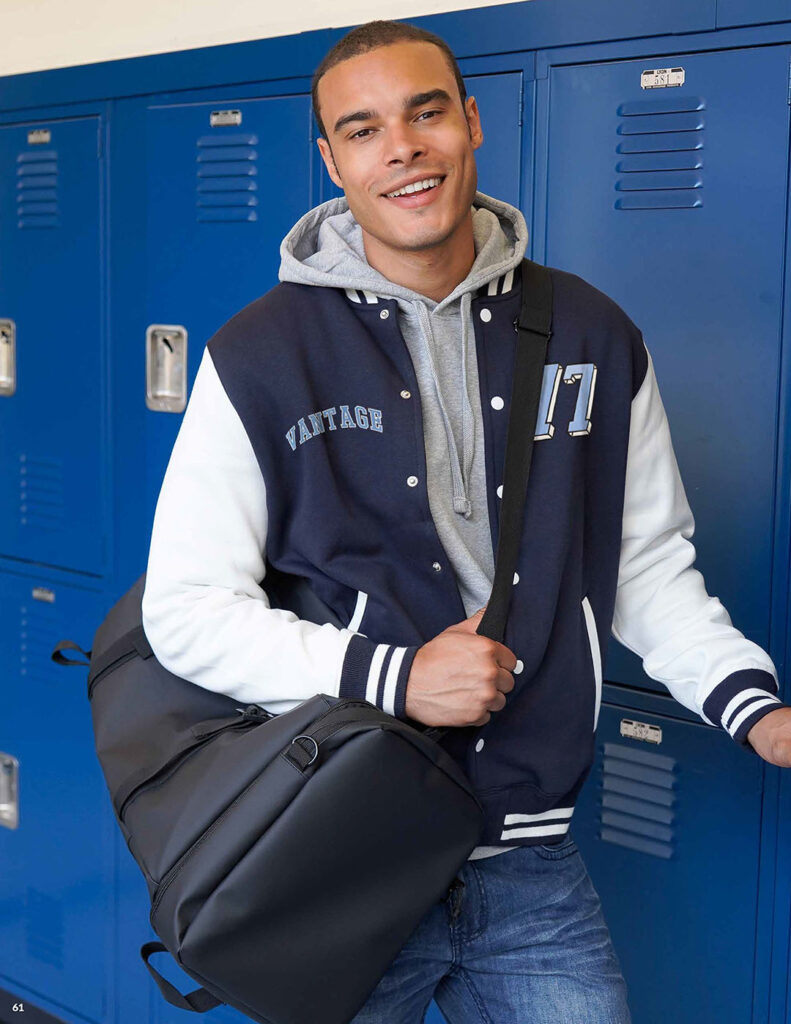 Person wearing a navy and white varsity jacket with a gray hoodie and carrying a black duffel bag in front of blue lockers.