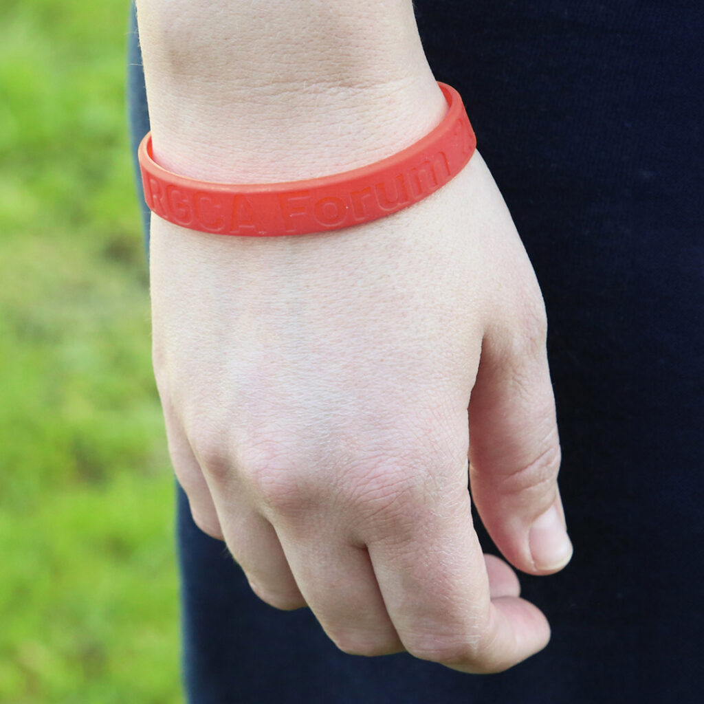 Close-up of a hand wearing a red silicone wristband with embossed text, symbolizing support for American Heart Month, against a blurred green outdoor background.