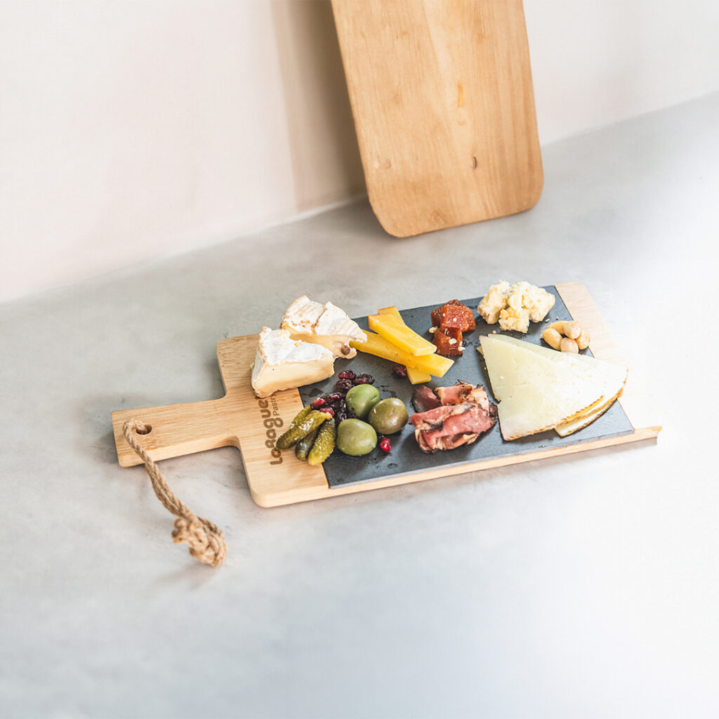 A wooden serving board topped with assorted charcuterie items, including cheeses, olives, pickles, sliced meat, and vegetables, arranged neatly on a slate surface.