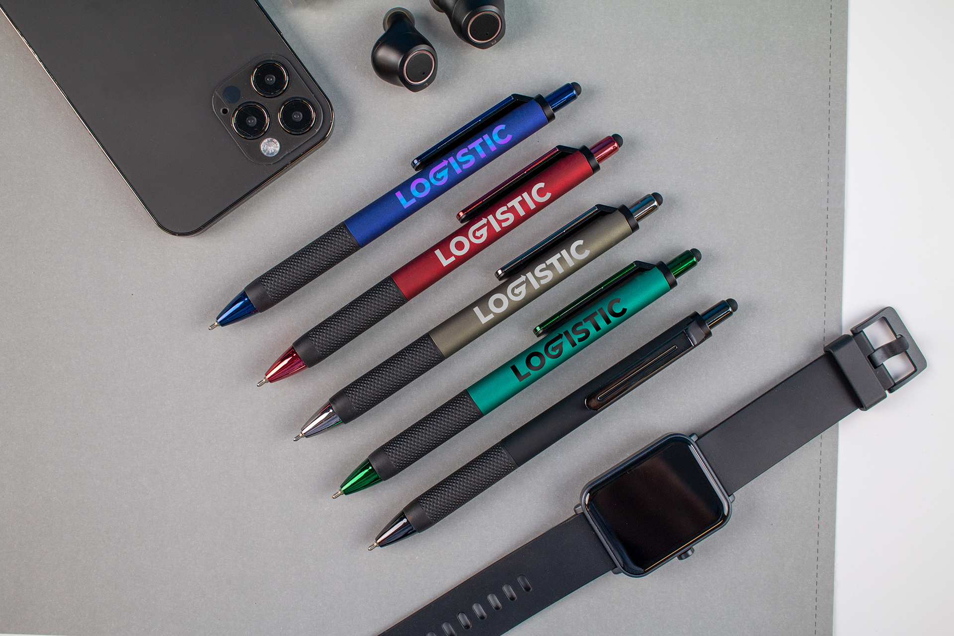 Flat lay of promotional items including five colored LOGISTIC‑branded pens, a smartphone, wireless earbuds, and a smartwatch arranged on a gray surface.