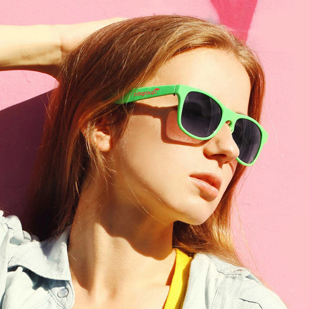 Person with long hair wearing a light denim shirt and neon green sunglasses, posing with one arm raised against a bright pink wall.