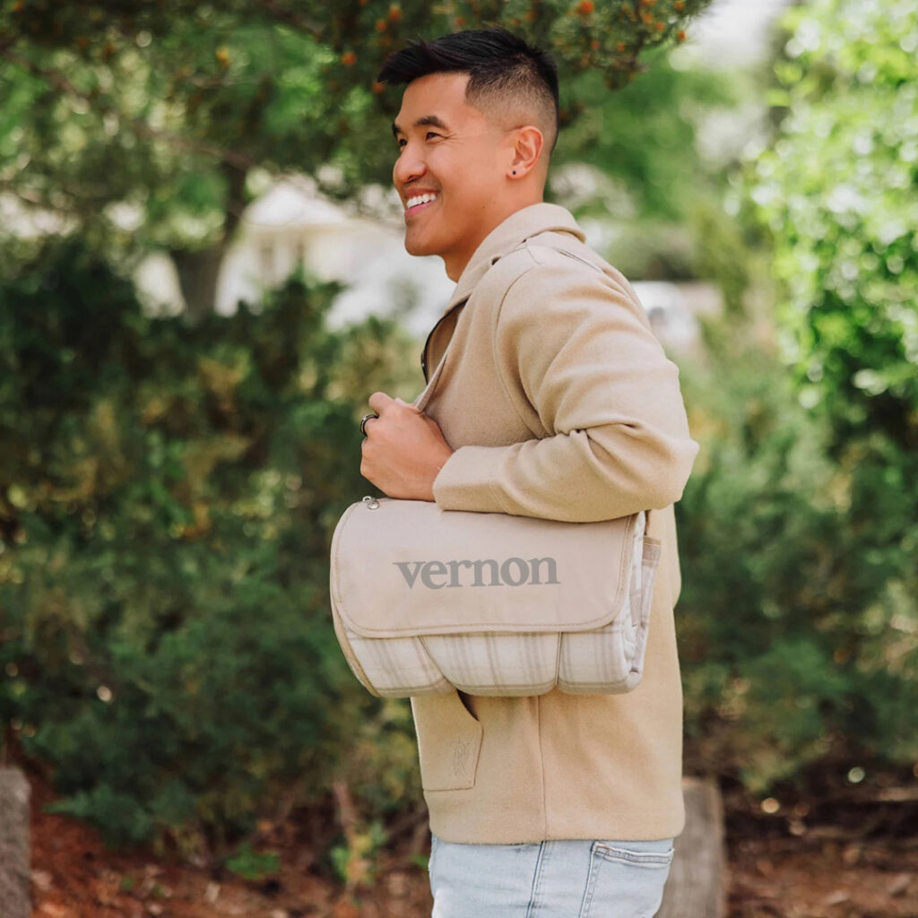 Person outdoors wearing a beige coat and carrying a light-colored roll-up picnic blanket, standing in front of green trees.