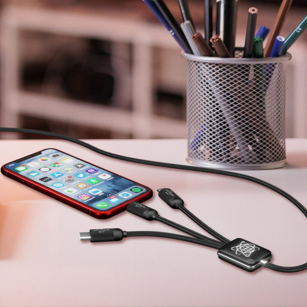 Multi‑device charging cable connected to a smartphone on a desk, with a metal cup of pens and pencils in the background.