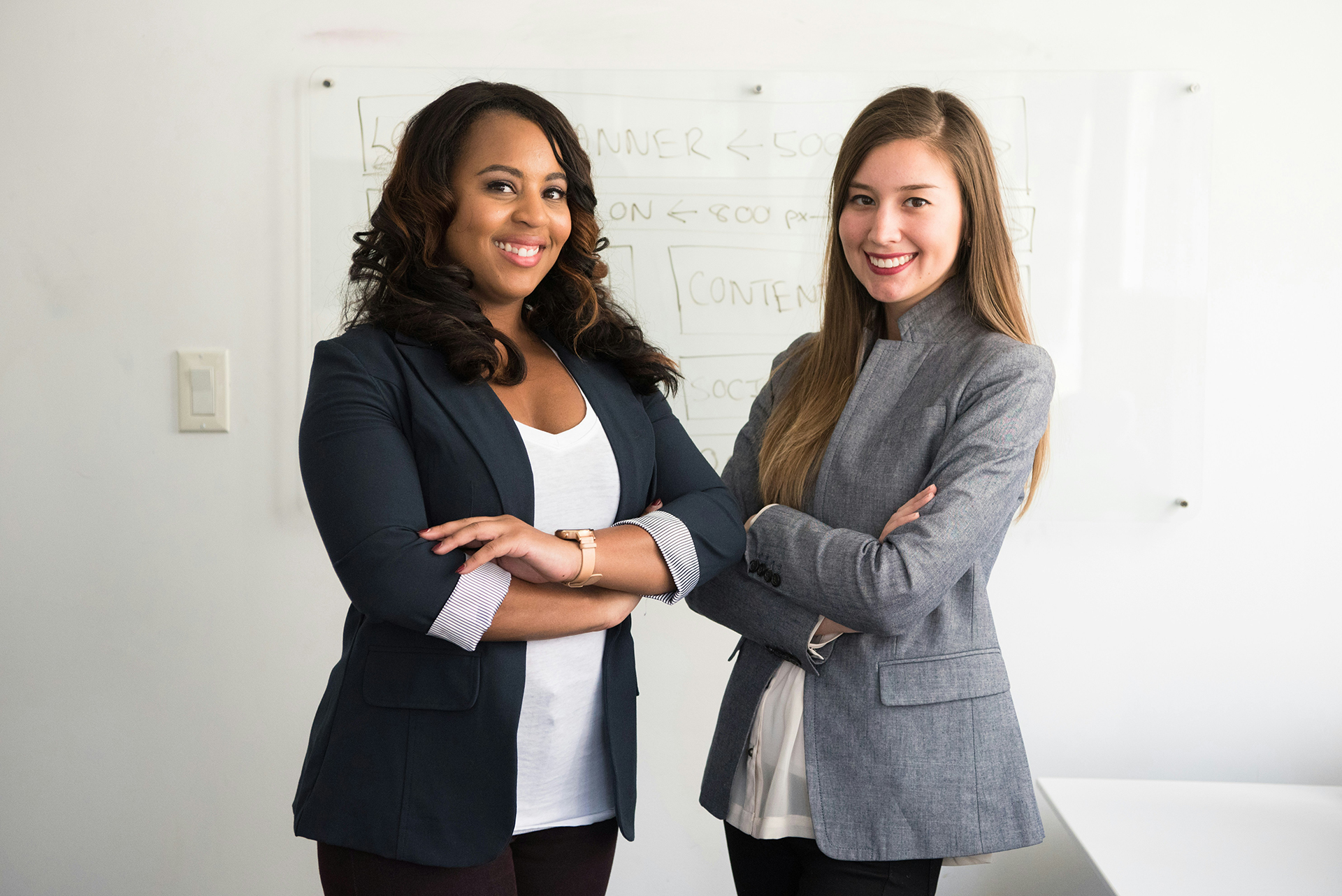 Two professionally dressed women standing with arms crossed in a bright office with a whiteboard in the background.