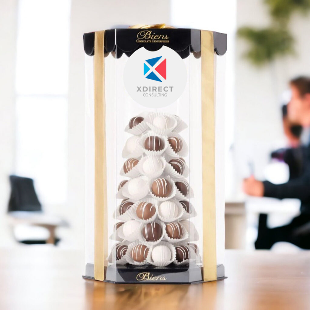 Tall clear gift tower filled with white and chocolate truffles, wrapped with gold ribbon and labeled with the XDirect Consulting logo, displayed on a table in a bright office setting.