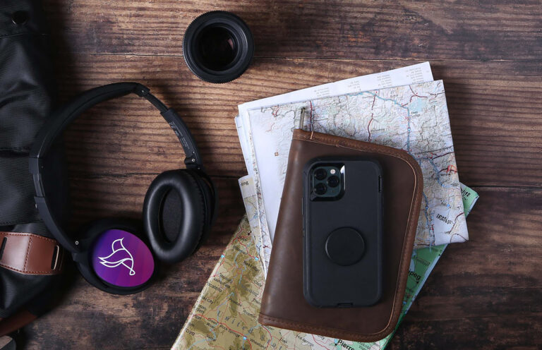 Flat lay of travel accessories on a wooden surface, including over‑ear headphones, maps, a phone in a case on a leather wallet, and a camera lens.