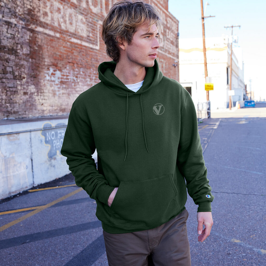 Person wearing a dark green hooded sweatshirt with a small logo on the chest, walking on a street with brick buildings in the background.