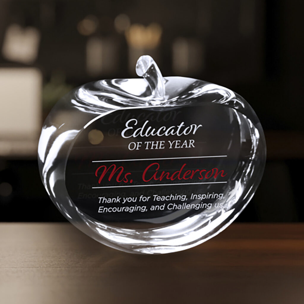Clear crystal apple award engraved with the text “Educator of the Year – Ms. Anderson” on a wooden surface.