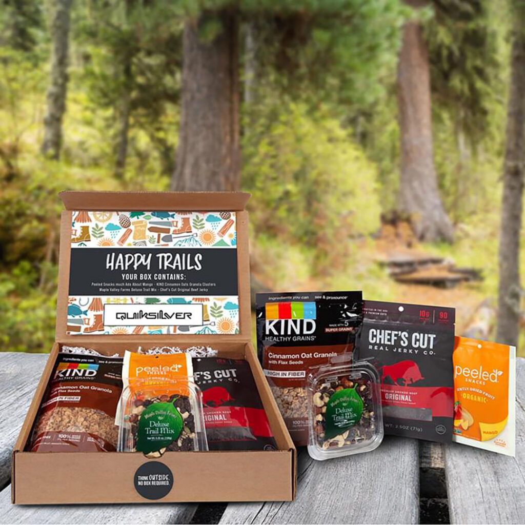 Open snack gift box with granola, trail mix, and jerky displayed on a wooden surface with a forest background.