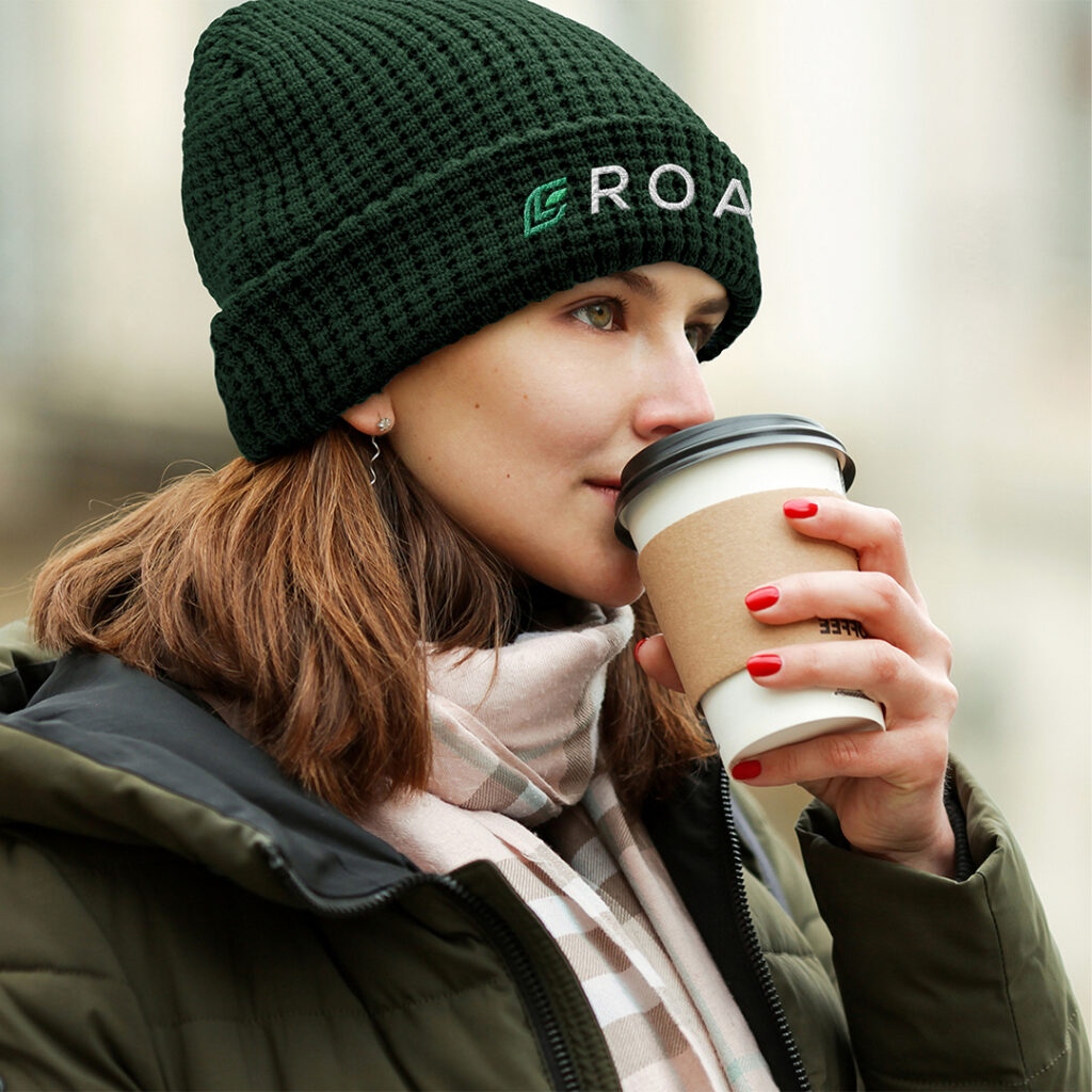 Person wearing a dark knitted beanie, winter coat, and plaid scarf while holding a takeaway coffee cup outdoors.