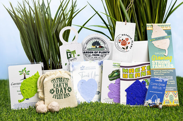 Eco‑friendly promotional items arranged on grass, including plantable seed paper shapes, Earth Day messaging, branded tags, and sustainability‑themed cards displayed in front of tall green plants.