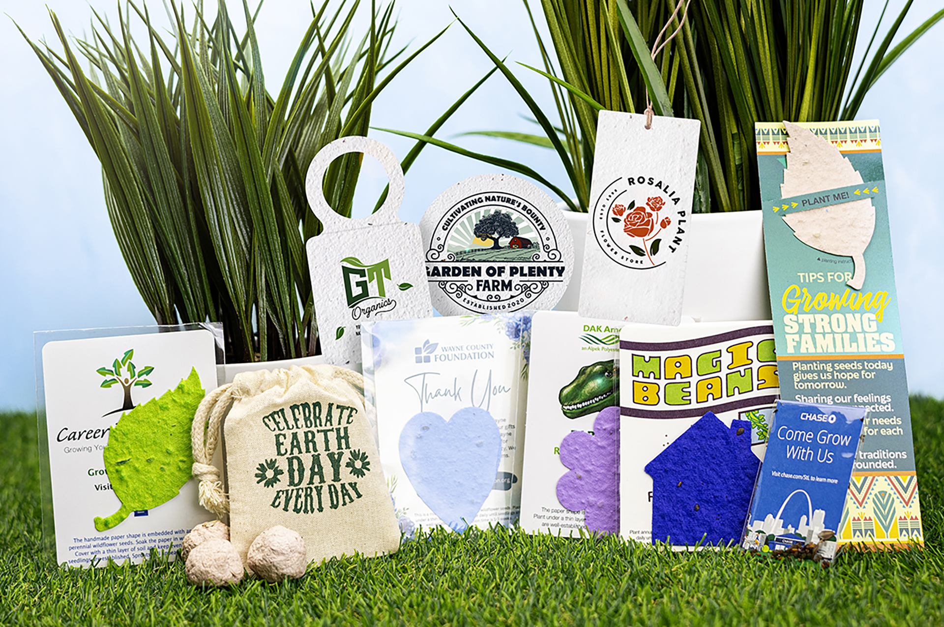 Eco‑friendly promotional items arranged on grass, including plantable seed paper shapes, Earth Day messaging, branded tags, and sustainability‑themed cards displayed in front of tall green plants.