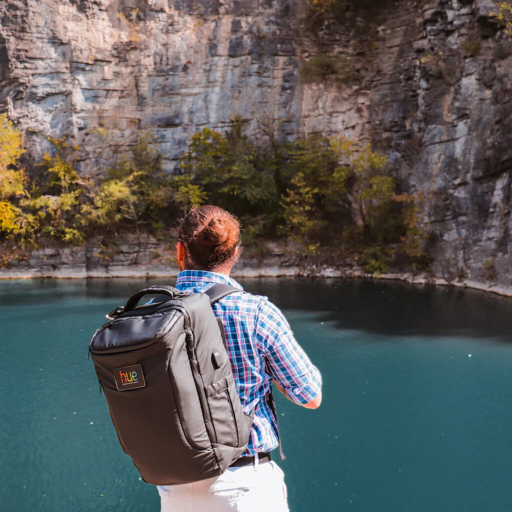 Person wearing a large black backpack standing at the edge of a blue lake, facing a tall rocky cliff with trees along the shoreline.