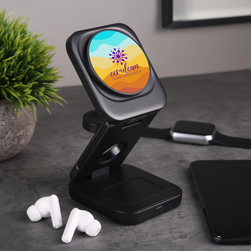 Black magnetic wireless charger displayed on a desk with earbuds, a smartwatch band, and a phone nearby; the charger screen shows a colorful ocean‑themed graphic.