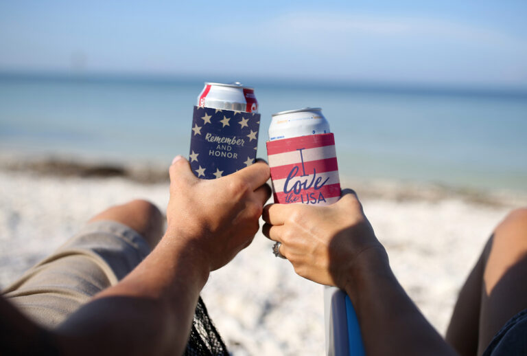 Two hands holding beverage cans in patriotic can sleeves with stars and stripes, resting over a sandy beach with ocean water in the background.