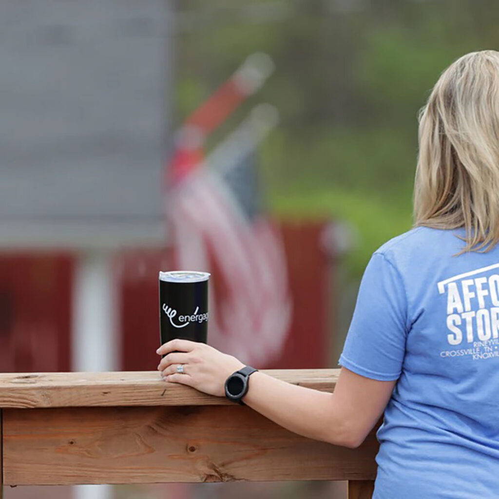 A person leaning on a wooden railing holding a black insulated tumbler, with an American flag blurred in the background.